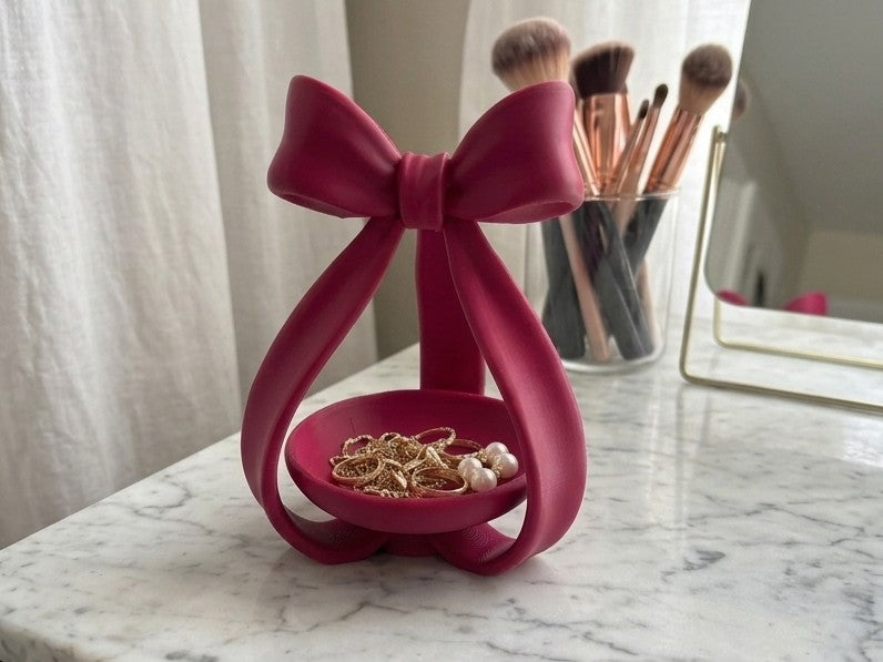 Tall coquette bow jewelry dish in pink holding gold rings and pearl earrings on a marble vanity